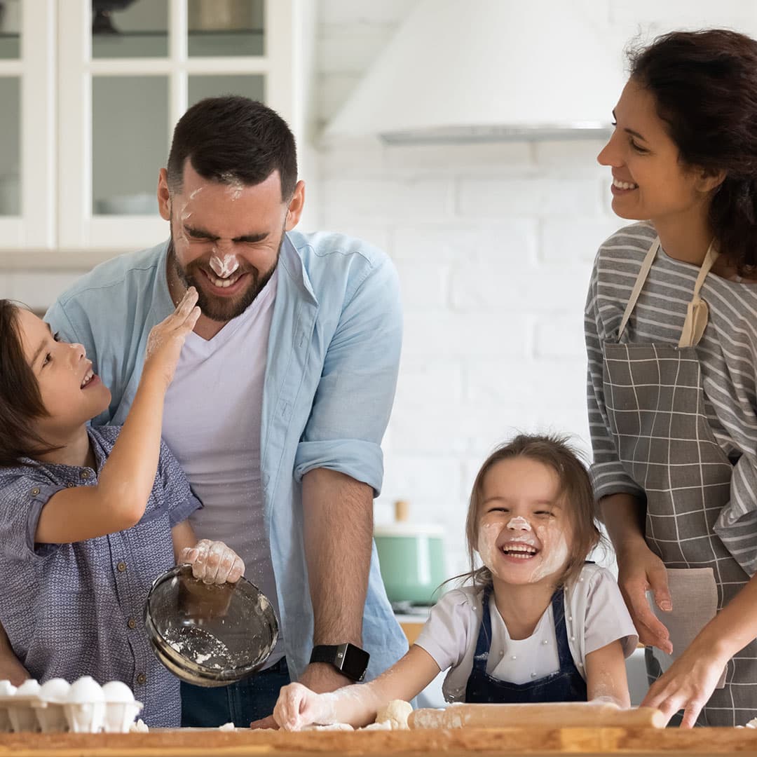 A family scene of baking together, with a little boy playfully throwing flour at his laughing father, a little girl smiling with flour on her face, and the mother beside her, smiling warmly.