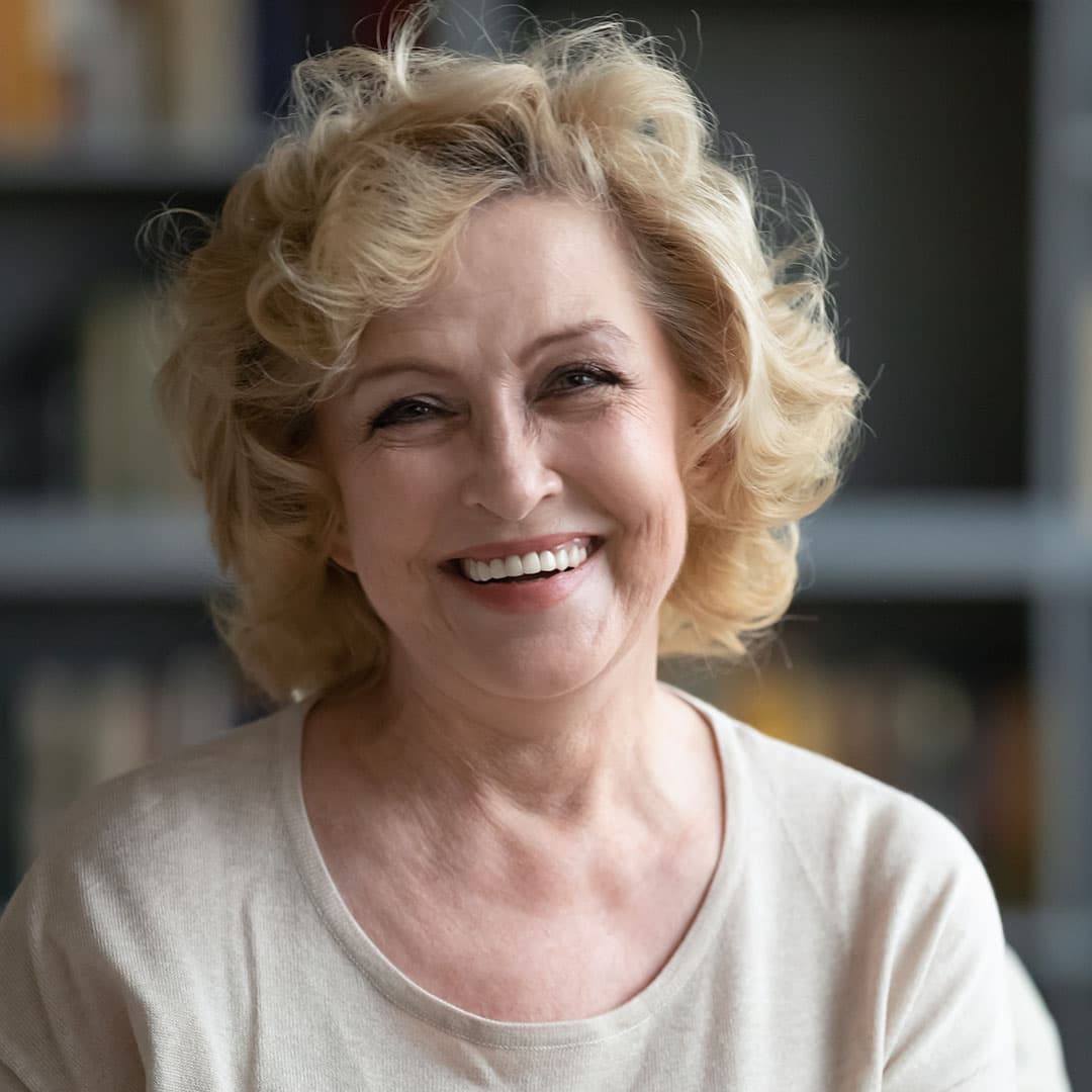 A close-up of an elderly woman with curly hair, smiling warmly while wearing a white shirt.