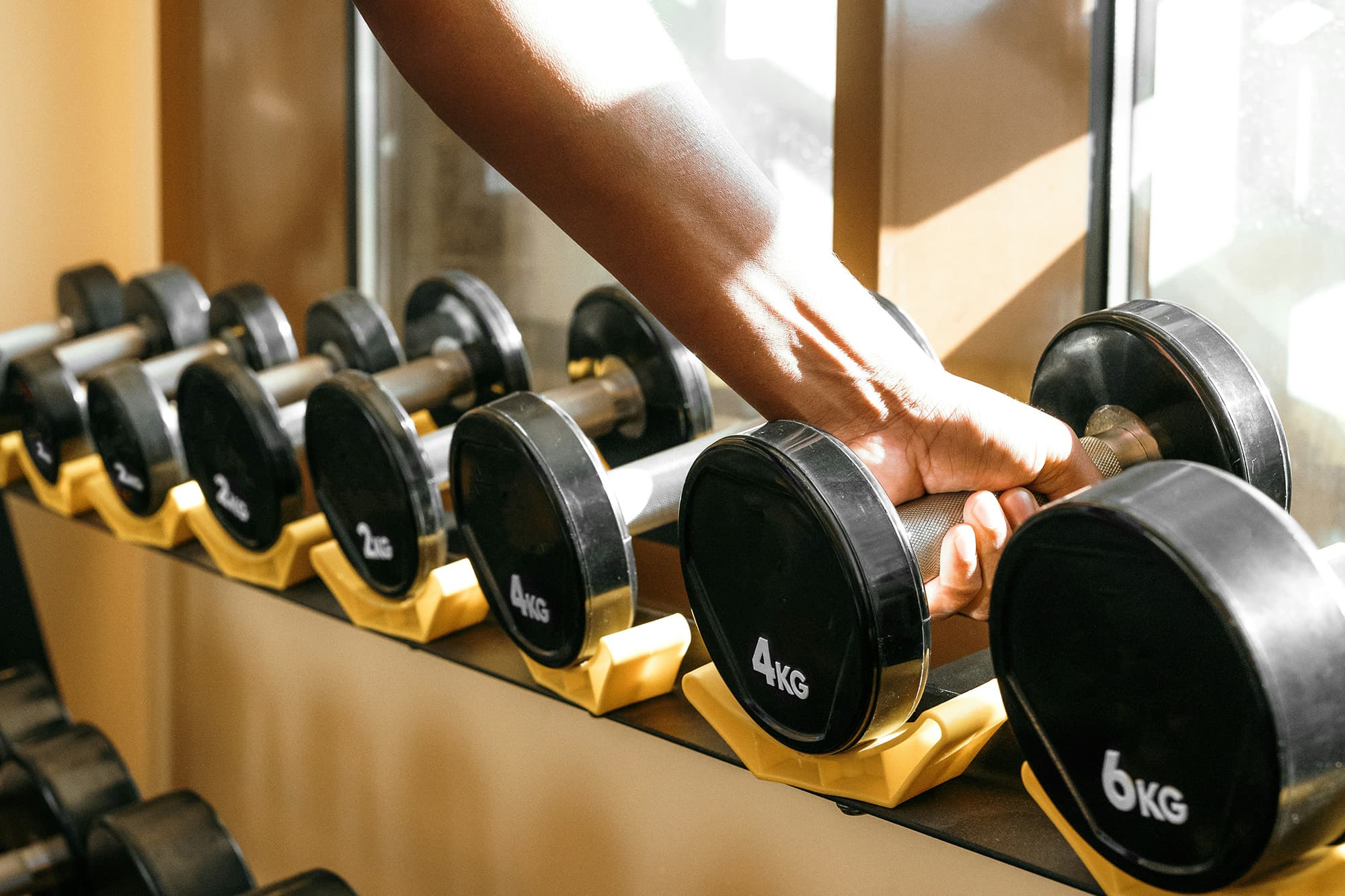 A close-up of dumbbells with different weights lined up in front of bright windows, with a hand reaching to grab the 4kg dumbbell.