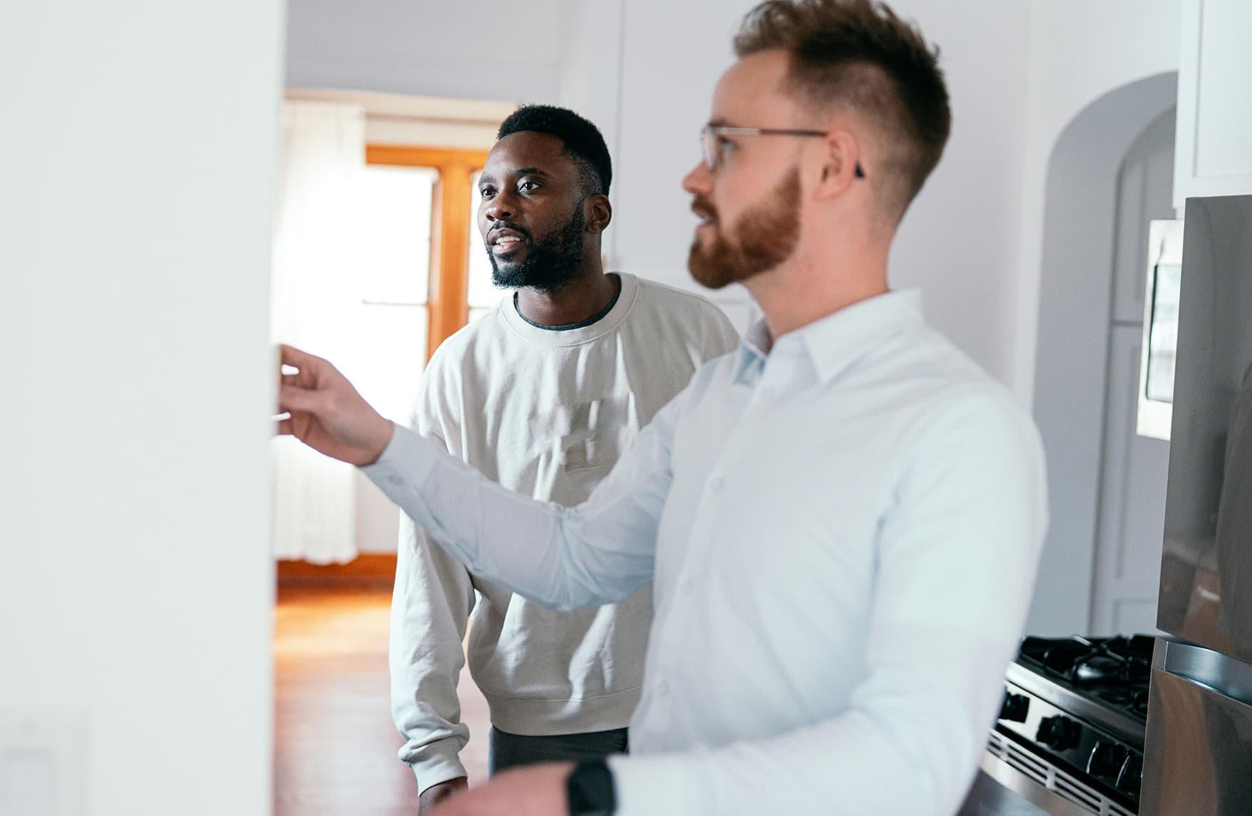 Two men dressed in white stand inside a house, looking thoughtfully at a wall, suggesting planning or renovation.