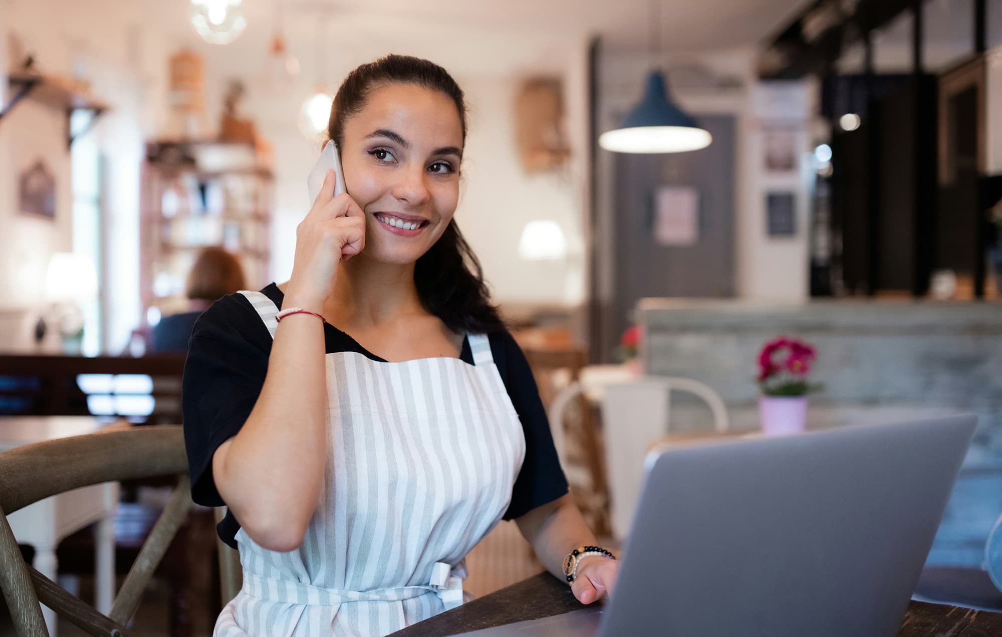 A person wearing a striped apron over a black T-shirt sits at a café table, smiling while talking on the phone, with an open laptop in front of them.