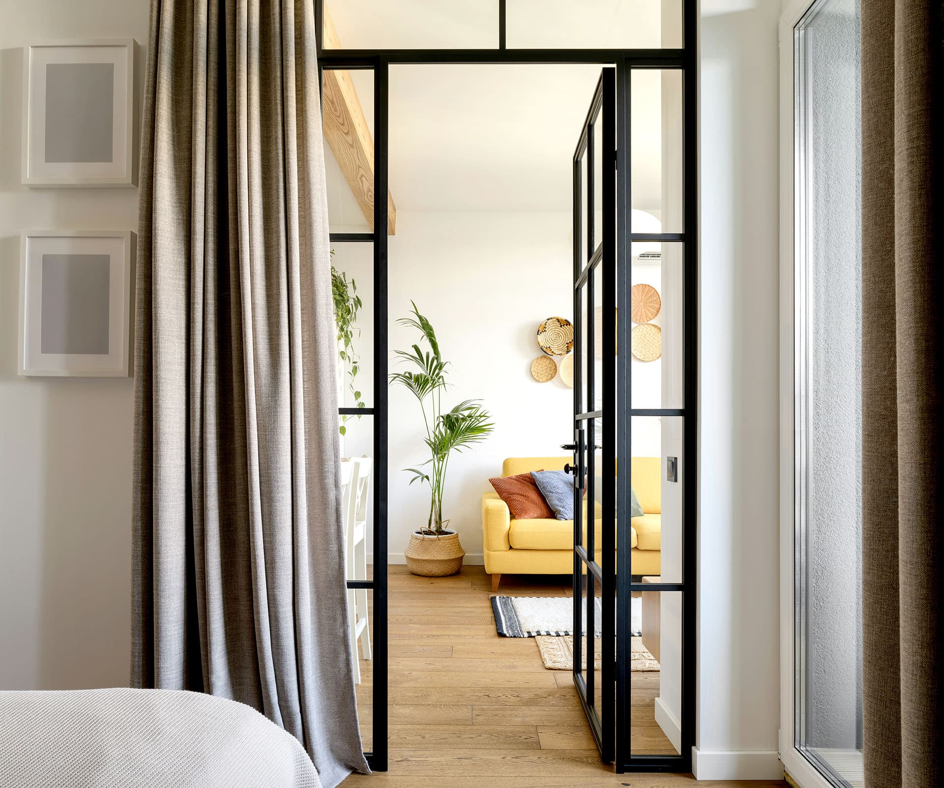 A view through a glass door with black frames into a room featuring a yellow sofa and a tall plant beside it.