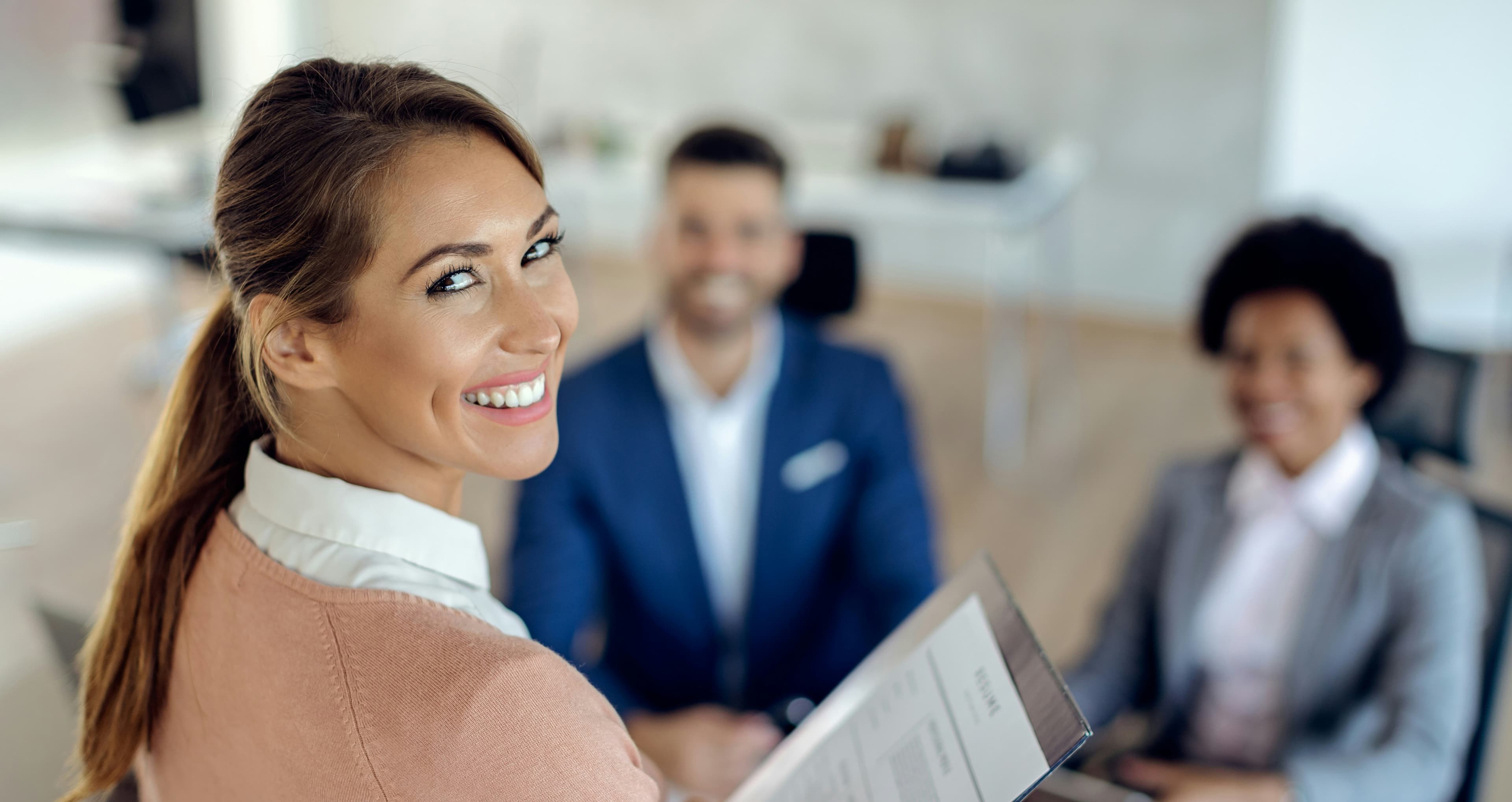 A woman in professional attire looks back smiling while holding her resume, with blurred figures in suits smiling in the background, suggesting an interview setting.
