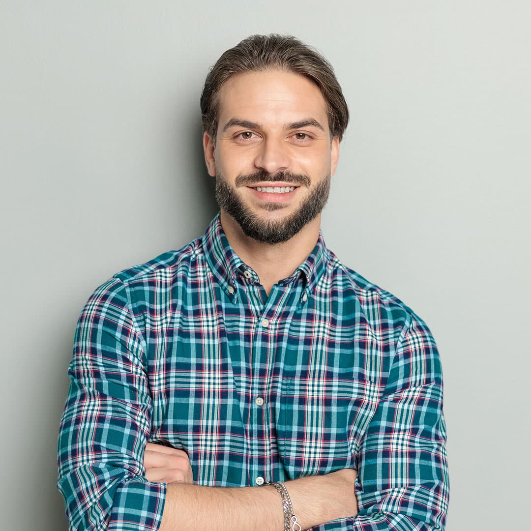 A bearded man wearing a plaid shirt stands confidently with his arms crossed, smiling, and wearing a silver bracelet on his wrist.
