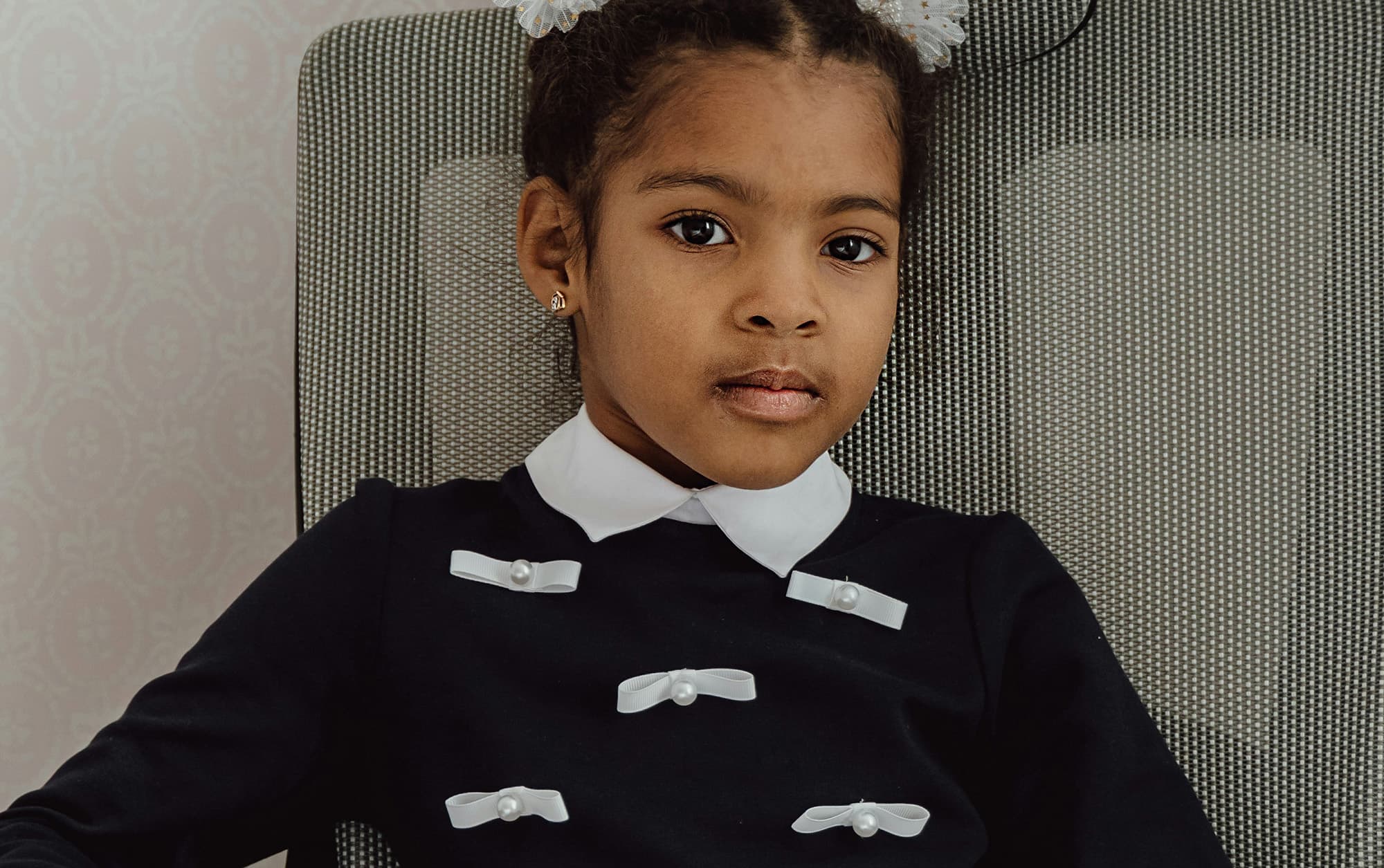 A close-up of a young girl sitting on a chair, her hair styled in two buns, wearing a black sweater with white ribbons all around.
