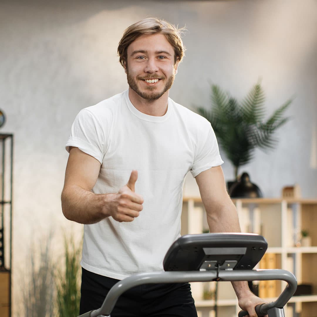 A man with blond hair and a beard giving a thumbs-up while working out on a treadmill, wearing a white T-shirt and smiling confidently.